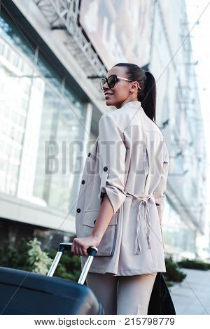 Enjoying her business trip. Rear view of beautiful young woman in sunglasses looking awaywith smile
while pulling her luggage outdoors.
