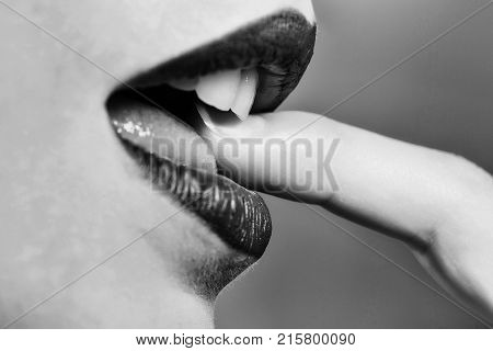 Female mouth with sexy purple lips and white teeth bites finger on grey wall