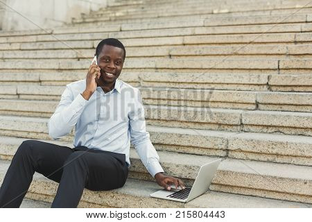 Happy black businessman working on laptop. African-american insuranse agent checking email on computer, talking on mobile, sitting on the stairs outdoors in urban area