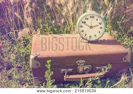 Vintage alarm clock on an old classic brown leather suitcase on a background of green grass. Retro toning. Travel concept
