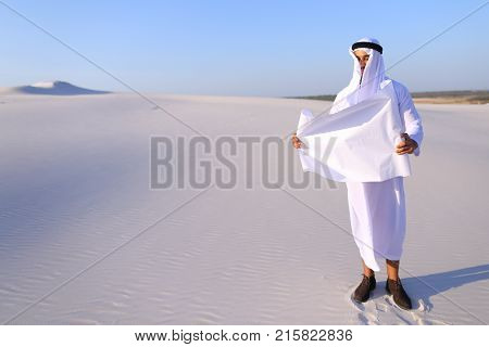 Beautiful Emirate Sheikh businessman studying project of future construction of complex, standing in middle of bottomless desert with white clean sand on clear warm day against blue sky. Swarthy Muslim with short dark hair dressed in kandura, long, spacio