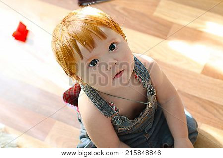 Close up portrait of cute little baby with ginger hair dressed in jeans overall sitting on the flloor and looking up
