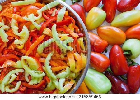 Juicy sliced lengthwise stripes of multi-colored peppers are lying in large round metal bowl on left and whole uncut peppers are lying on table on right.