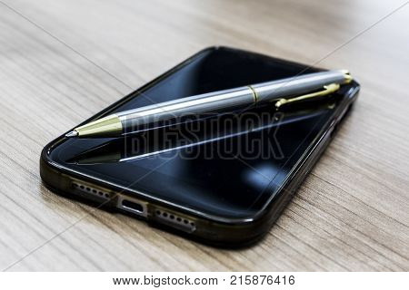 Gold-silver ballpoint pen on Black mobile phone and on wood background. Modern with a businessman.