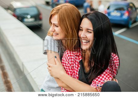 Two atractive young girls hugging, laughting and sitting near road in the street. one brunette girl in red plaid shirt, another redhead girl in gray shirt and blue skirt. concept of sincere friendship