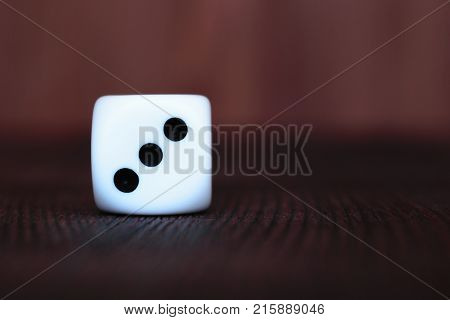 Single white plastic dice on brown wooden board background. Six side cube with black dots. Number 3.