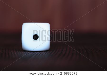 Single white plastic dice on brown wooden board background. Six side cube with black dots. Number 1.