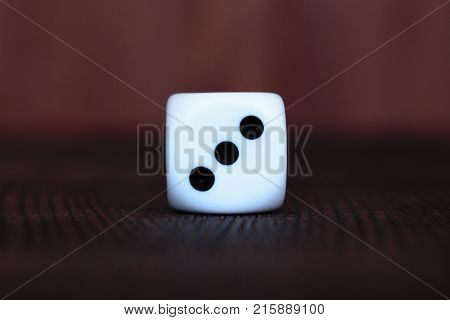 Single white plastic dice on brown wooden board background. Six side cube with black dots. Number three.