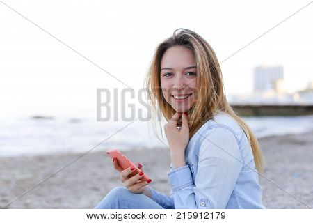 Cute girl holding cell phone and leads in social network or post photos, laughing and smiling, sharing impressions of beauty of sea, sitting on sandy beach on backdrop of sea and sky on clear evening . Young European-looking woman with long blond hair dre