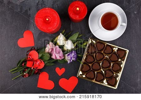 A cup of tea on a stand with a box of chocolates and burning candles, a bouquet of Japanese roses and carved hearts on a stone background. View from above