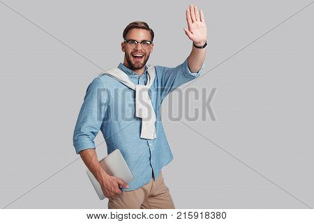 Hello! Good looking young man in eyeglasses holding his digital tablet and gesturing while standing against grey background