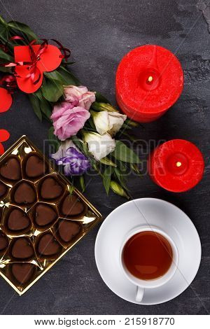 A cup of tea on a stand with sweets, with a box of sweets showing not completely, a bouquet of Japanese roses and burning candles on a stone background. View from above