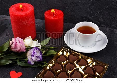 A cup of tea on a stand next to a box of chocolates. A bouquet of Japanese roses and burning roses on a stone background