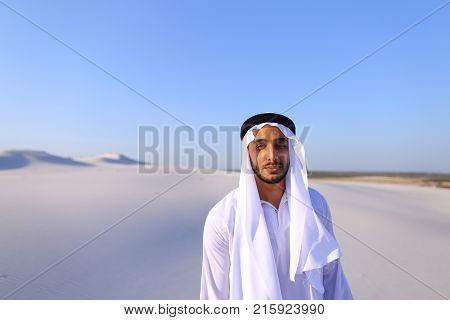 Cheerful Muslim guy walks along white sand of wide desert on warm summer evening. Arab young man in national clothes dances on spot and laughs giggly, adjusting hands with harness in wind. Swarthy, handsome Muslim with short dark hair dressed in kandura, 