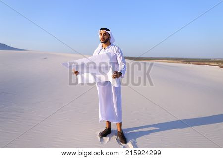 Beautiful Emirate Sheikh businessman studying project of future construction of complex, standing in middle of bottomless desert with white clean sand on clear warm day against blue sky. Swarthy Muslim with short dark hair dressed in kandura, long, spacio