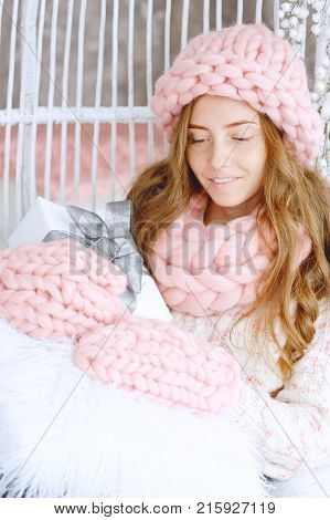 Young woman with red hair and dressed in sweater and knitted pink hat and snood sitting in her favourite chair holding holiday gifts