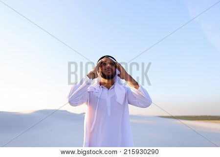 Portrait of tired Arab man, who holds head and feels migraine, tries to calm pain and massages whiskey, standing in middle of bottomless desert with snow-white sand on sunny summer day. Swarthy Muslim with short dark hair dressed in kandura, long, spaciou