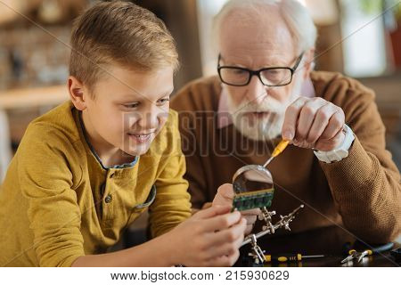 Young engineer. Joyful smart nice boy holding a microscheme and looking at it while using magnifying glass