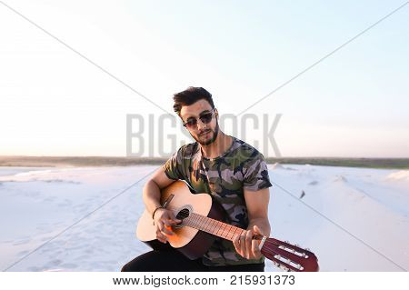 Handsome Arabian guy plays tune on guitar, drives strings and sings song, sitting on hill in middle of wide sandy desert on warm summer evening at sunset. Swarthy man with dark hair and short haircut in sunglasses dressed in camouflage t-shirt, black pant