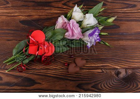 A bouquet of Japanese roses tied with a ribbon with a heart next to candy in the form of hearts and a beak lie on a wooden background. View from above...