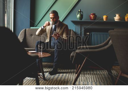 Middle-aged Businessman Drinking Coffee And Uses Mobile Phone In The Loby