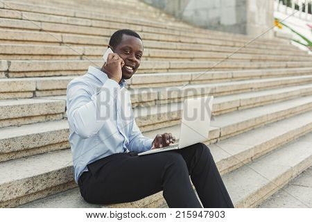 Happy black businessman working on laptop. African-american insuranse agent checking email on computer, talking on mobile, sitting on the stairs outdoors in urban area