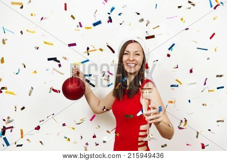 Beautiful smiling caucasian young happy woman in red dress and Christmas hat standing with big tree toy and glass of champagne on white background confetti. Santa girl isolated. New Year 2018 holiday.