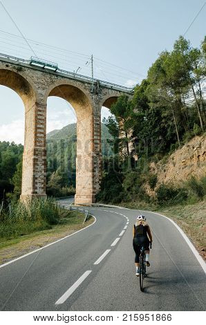 Strong and empowered professional female cyclist explores road less ridden on carbon bicycle rides in european country side on cycling vacation in summer under ancient bridge