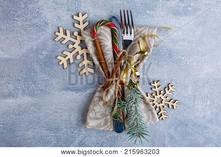 On a worn blue surface, the knife and fork are wrapped in a serving napkin and decorated with a golden ribbon, fragrant cinnamon sticks, Christmas candy and small twigs of a Christmas tree, beside wooden decorative snowflakes. View from above.