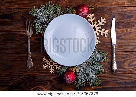 On the wooden table an empty white round plate, a knife and a fork are decorated with branches of a Christmas tree with red balls. View from above.