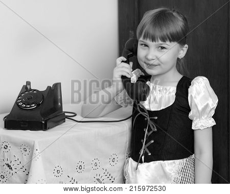 A sweet little blonde with long hair braided in a bun and a short bangs on her head. In a beautiful dress with a white apron.The girl dials the number on the old black phone.