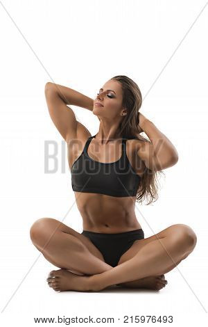 Muscular female bodybuilder wearing black top and pants with her dark long hair loose sitting in a yoga pose isolated on white background