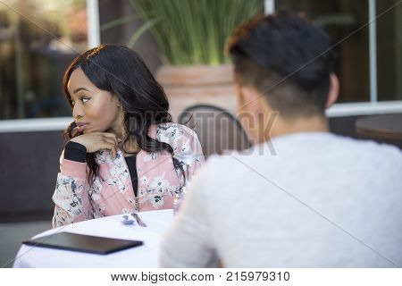 Black female disinterested with a blind date at an outdoor restaurant. They are sitting and chatting like speed dating. The image depicts relationship problems.