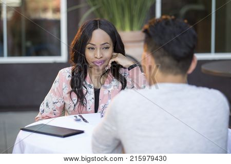 Black female disinterested with a blind date at an outdoor restaurant. They are sitting and chatting like speed dating. The image depicts relationship problems.
