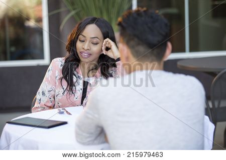 Black female disinterested with a blind date at an outdoor restaurant. They are sitting and chatting like speed dating. The image depicts relationship problems.