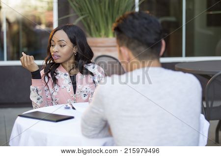 Black female disinterested with a blind date at an outdoor restaurant. They are sitting and chatting like speed dating. The image depicts relationship problems.