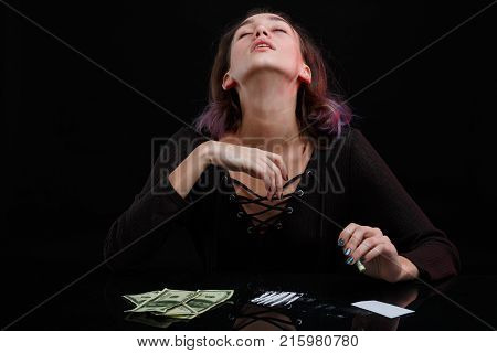 A young girl junkie throws her head back, kicks off drugs, next to the table is dosed cocaine and dollar bills. On a black background.
