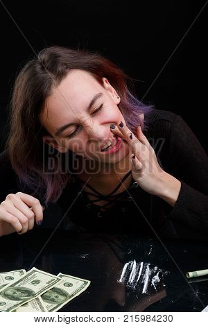 A young girl junkie rubs a white powder of cocaine into the gum, next to the dosed cocaine and a few dollar bills. On a black background.