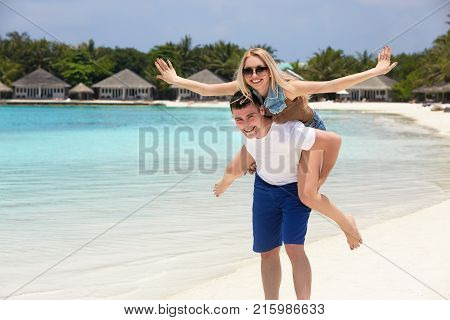 Portrait of smiling man carrying woman on his back along the sea shore. Guy giving piggyback ride to girlfriend by blue ocean. Young family having fun on tropical resort beach.