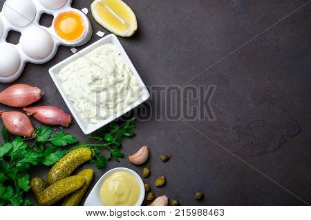 Homemade traditional french sauce remoulade in a white bowl with ingredients lemon eggs shallot pickles parsley mustard on a black dark stone slate background. Top view horizontal copy space