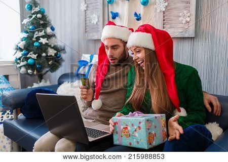 Happy young couple smiling while shopping online, on New Year's Eve, sitting on sofa using credit card and laptop, girl holding gift box in hands.