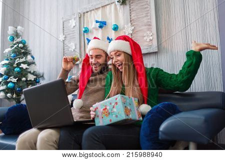 Happy young couple smiling, talking on video communication, making an online purchase, on New Year's Eve, sitting on sofa using credit card and laptop, girl holding gift box in hands.