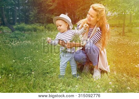 Happy Mother With Her Little Son In A Big Hat Are Walking On A Green Meadow In A Summer Park With A 