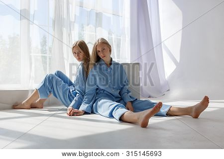 Two Young Pretty Twin Girls In Identical Blue Suits With Long Blond Hair Sitting On White Cyclorama 