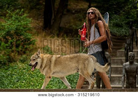 Prague, Czech Republic - August 1, 2019: White Man With Dreadlocks And With Dog On A Leash. In The B