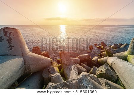 Beautiful Sunset Seascape. Breakwaters Tetrapods Ashore Of Pier. Cargo Ships On The Horizon. Travel 