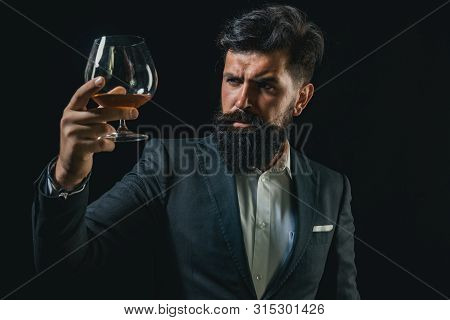 Degustation Of Elite Alcohol. Confident Bearded Man In Black Suit With Glass Of Cognac. Man With Bea
