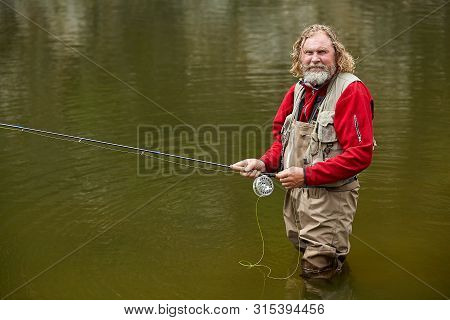 Mature Bearded White Man In Waterproof Clothing With Fishing Rod In His Hand Is Standing In River An