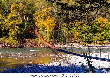 Narrow Metal Foot Bridge Across Mountain River In Autumn.