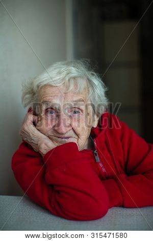 Very old woman in her house, close-up portrait sitting at a table in a red jacket. Plight of pensioners.